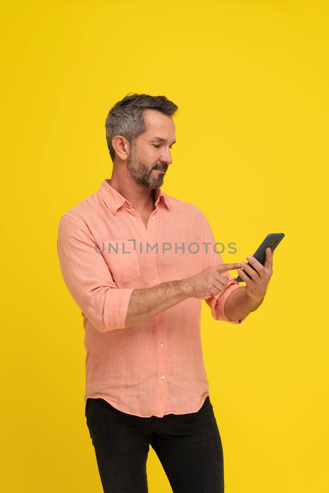 Masculine, fit middle aged grey haired handsome man holding smartphone looking at it wearing peach shirt isolated on yellow. Mature muscled man with phone, mobile app advertisement.