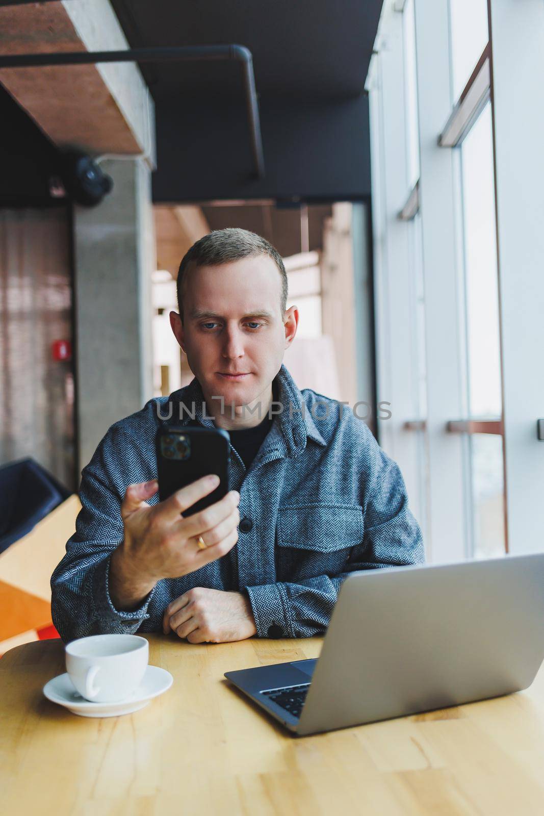 Smiling male entrepreneur in casual wear sitting at table with laptop and talking on smartphone in modern workspace with large window during day