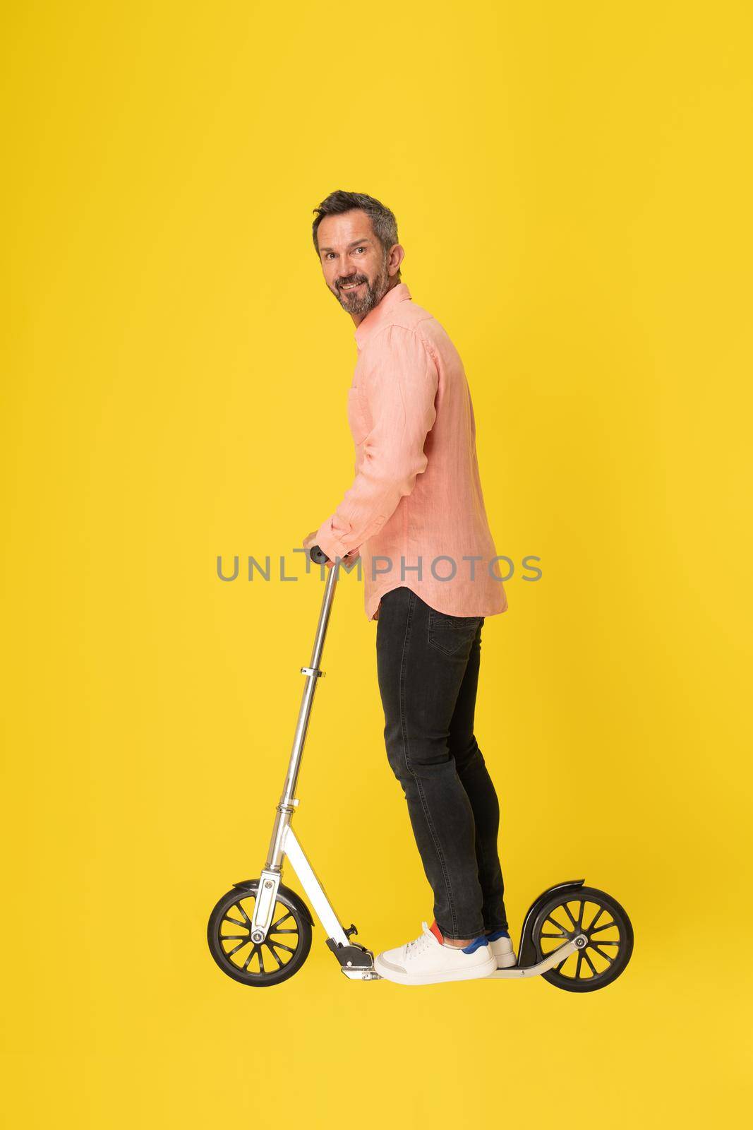Middle aged grey haired man ride scooter standing sideways looking at camera wearing peach shirt and black jeans isolated on yellow background. Fit masculine handsome mature businessman on the go.