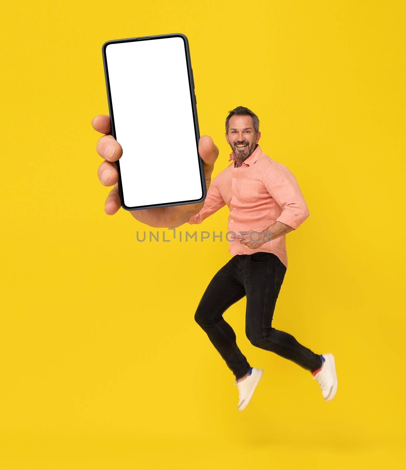 Excited middle aged man 40s jump with smartphone with white screen in hand happy smiling on camera wearing peach shirt and black jeans isolated on yellow background. Mobile app advertisement.