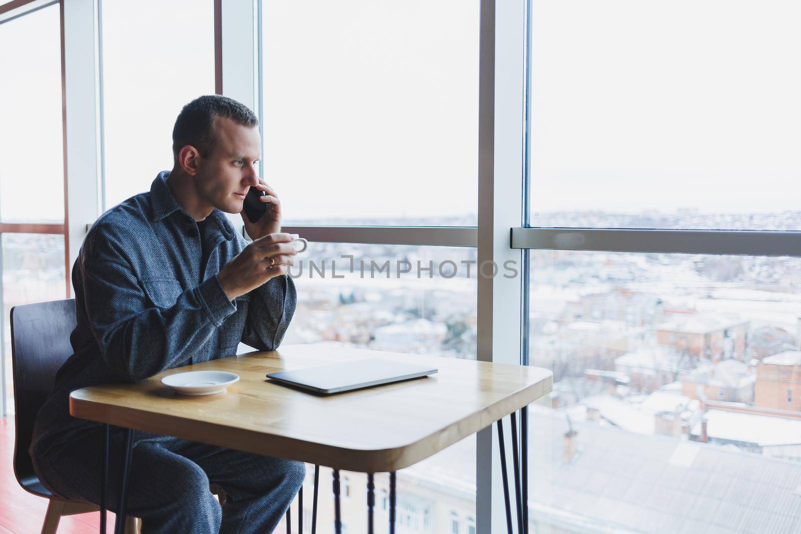 Happy man with coffee calling via smartphone while remote working laptop at work desk, cheerful freelancer enjoying coffee break while working remotely