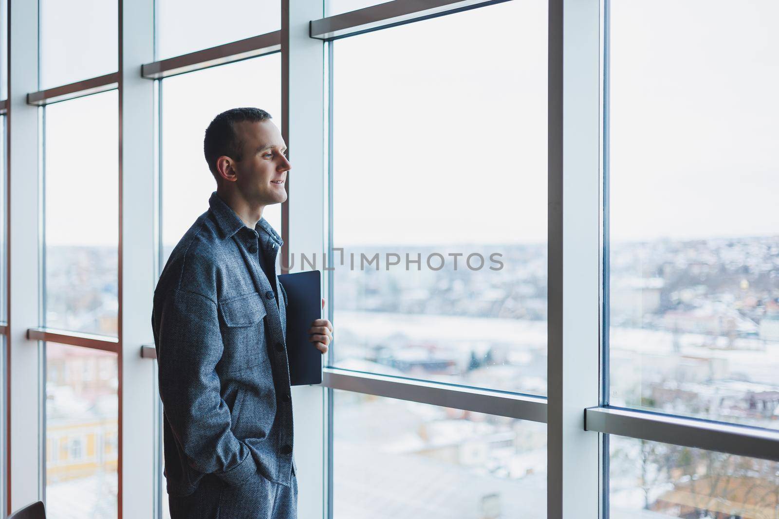 Portrait of a smart intelligent male manager, he is holding a laptop for research in the office, a business man in elegant clothes playing on the touchpad. Standing in the office by a large window