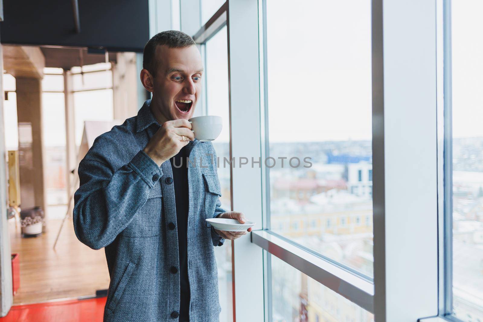 A young man of European appearance in a casual suit drinks coffee and looks out the window