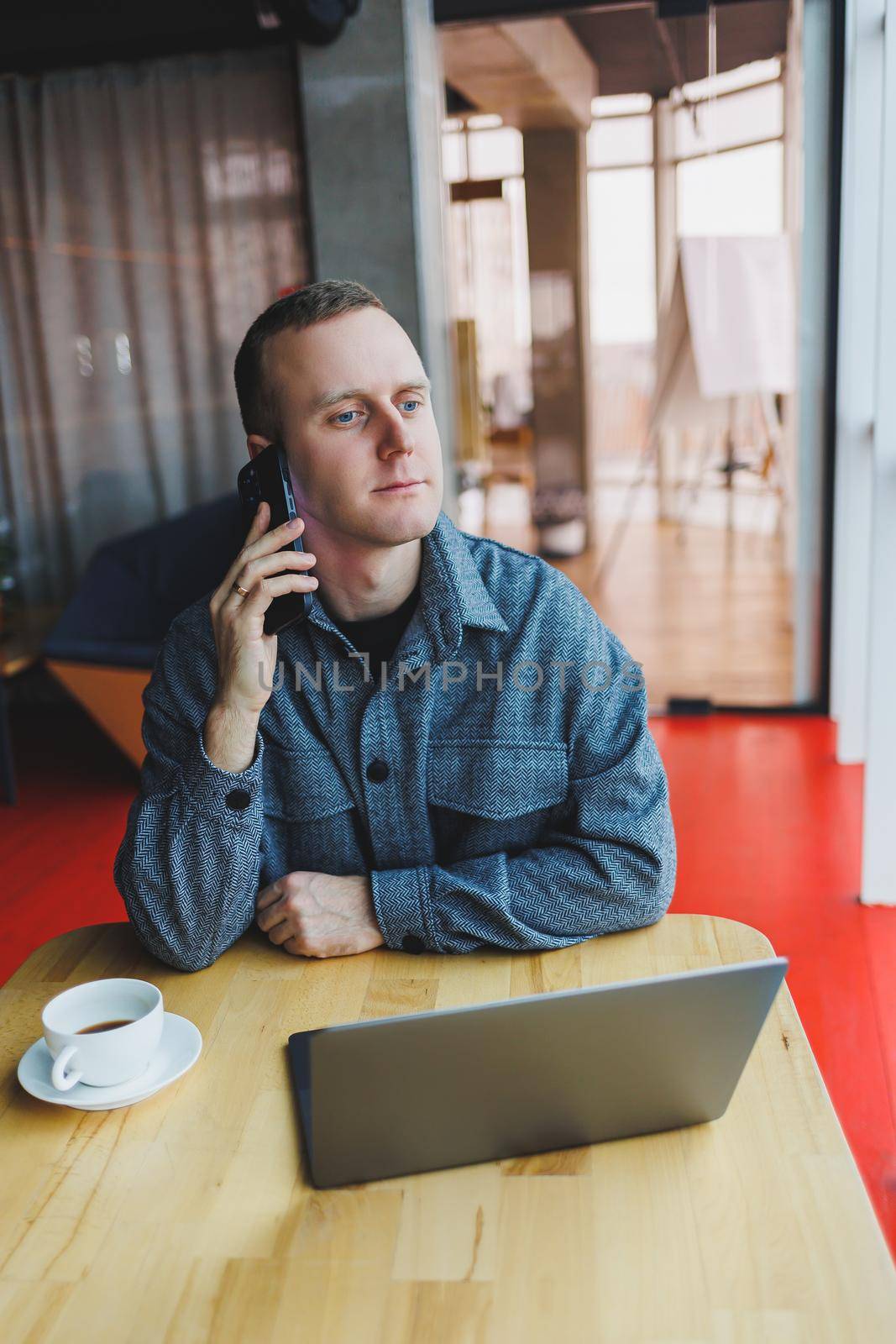 Smiling male entrepreneur in casual wear sitting at table with laptop and talking on smartphone in modern workspace with large window during day