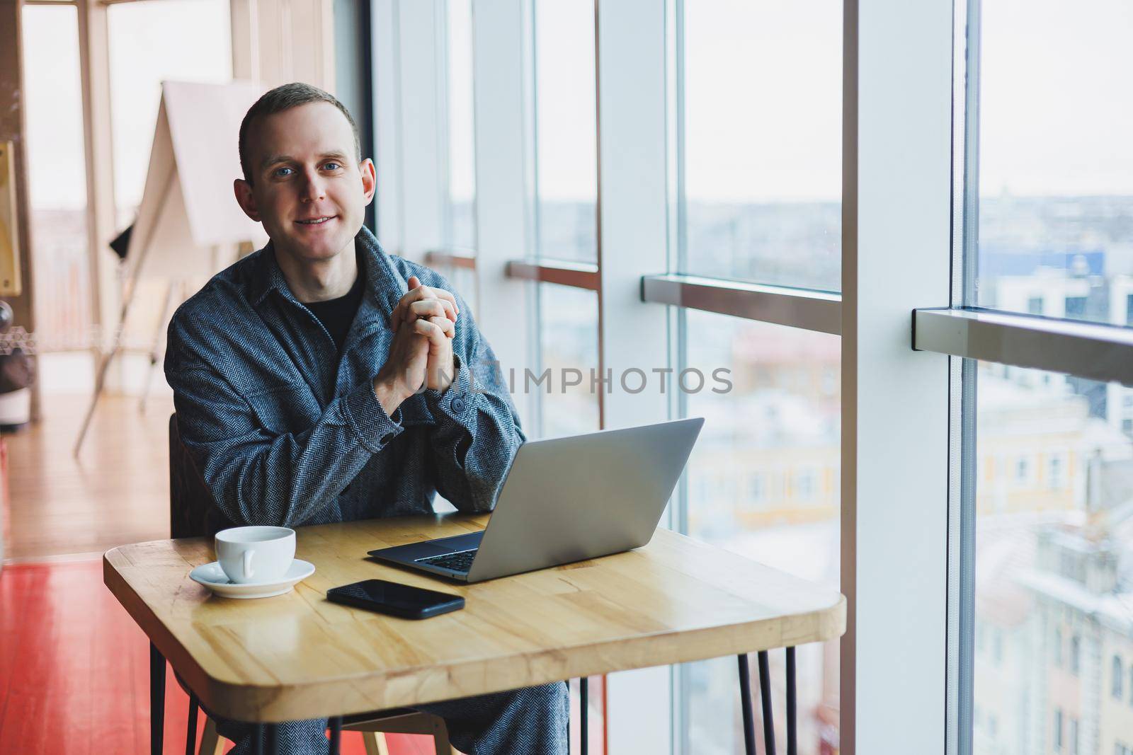 Successful happy business man is sitting at a table in a cafe, holding a cup of coffee and using a laptop.