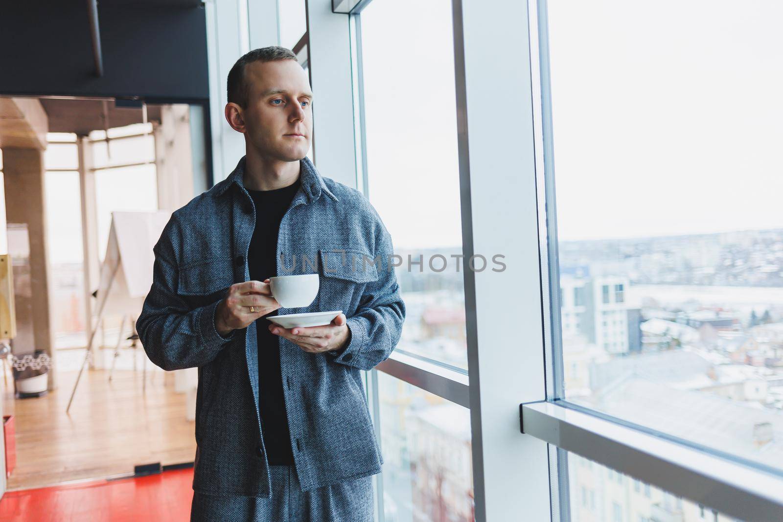 A young man of European appearance in a casual suit drinks coffee and looks out the window