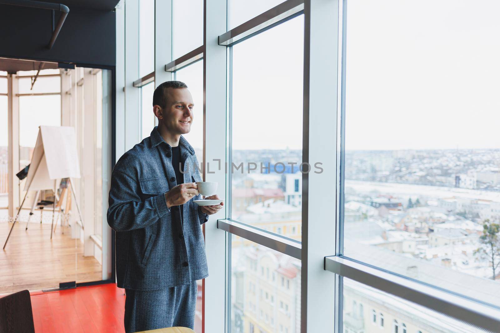 A young man of European appearance in a casual suit drinks coffee and looks out the window