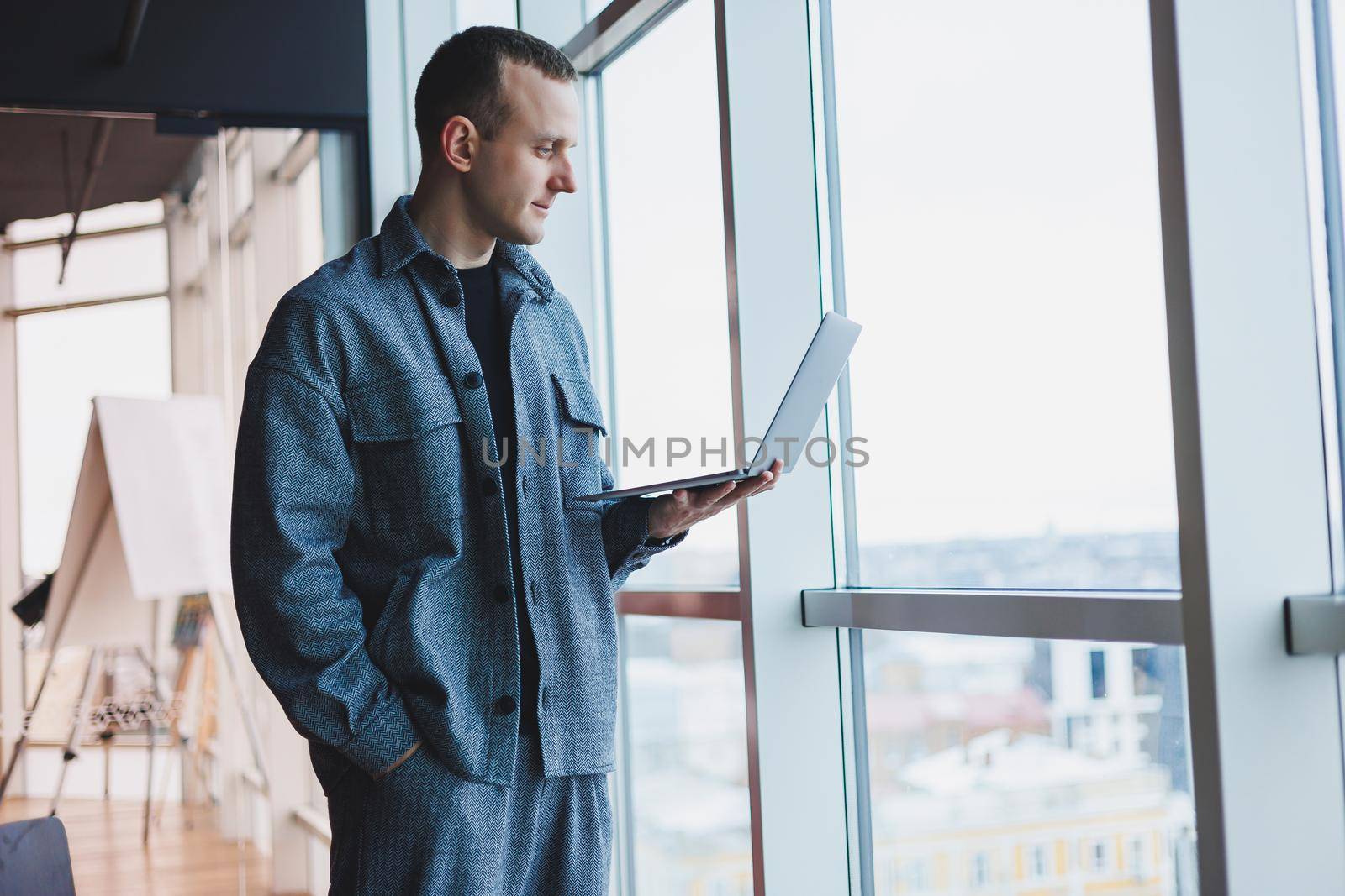 Portrait of a smart intelligent male manager, he is holding a laptop for research in the office, a business man in elegant clothes playing on the touchpad. Standing in the office by a large window