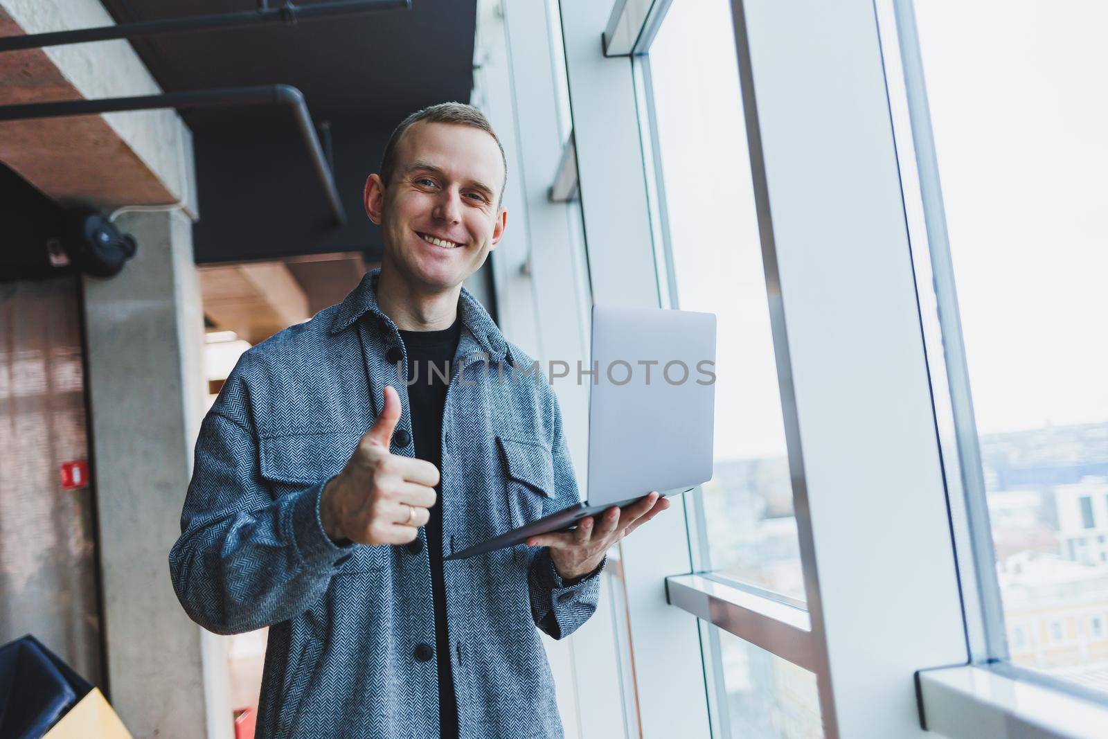 Portrait of a smart intelligent male manager, he is holding a laptop for research in the office, a business man in elegant clothes playing on the touchpad. Standing in the office by a large window