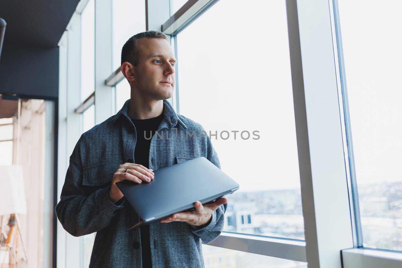 Portrait of a smart intelligent male manager, he is holding a laptop for research in the office, a business man in elegant clothes playing on the touchpad. Standing in the office by a large window