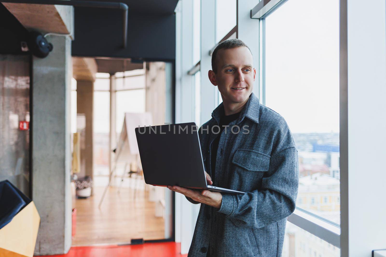 Portrait of a smart intelligent male manager, he is holding a laptop for research in the office, a business man in elegant clothes playing on the touchpad. Standing in the office by a large window