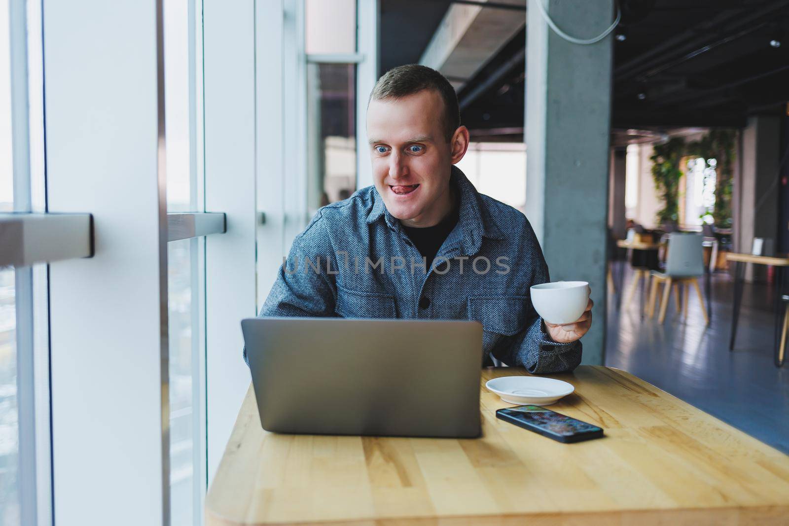 Successful happy business man is sitting at a table in a cafe, holding a cup of coffee and using a laptop.