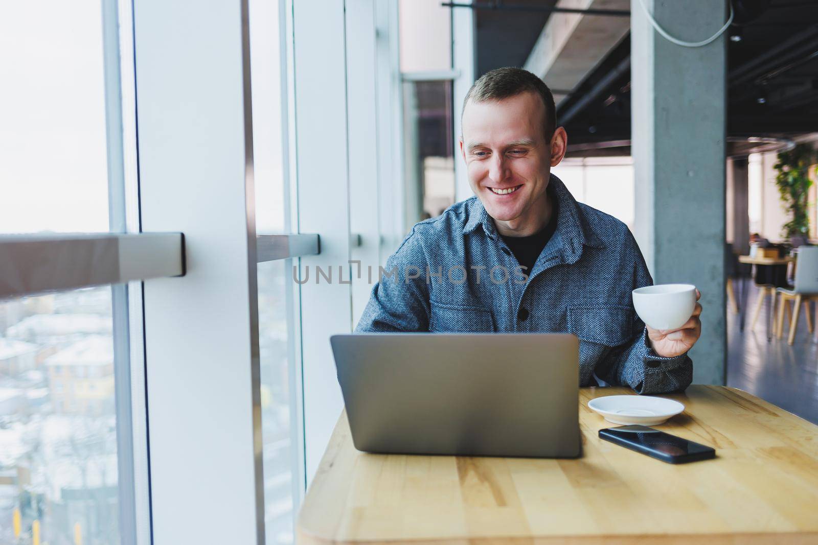 Successful happy business man is sitting at a table in a cafe, holding a cup of coffee and using a laptop.