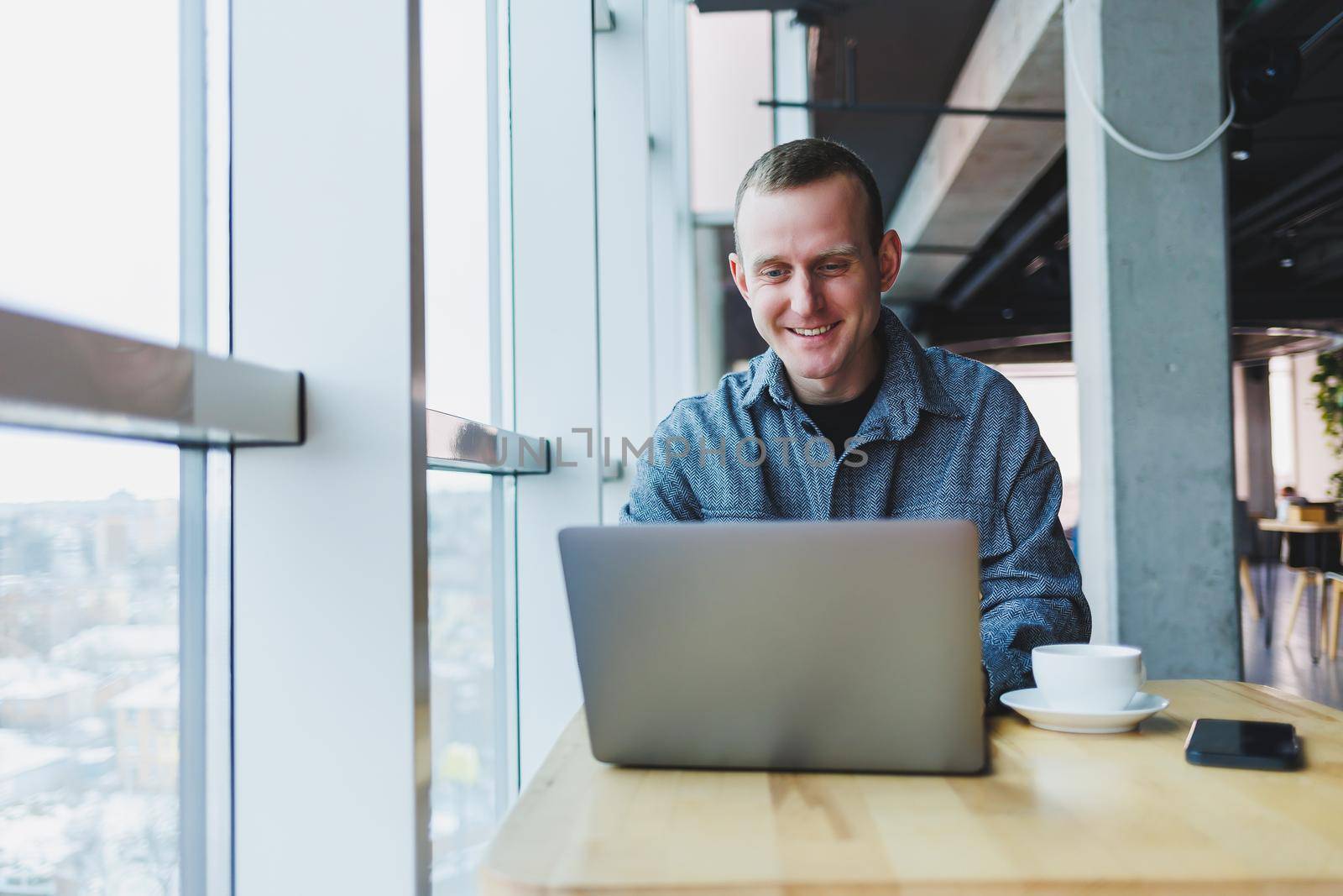 Successful happy business man is sitting at a table in a cafe, holding a cup of coffee and using a laptop.