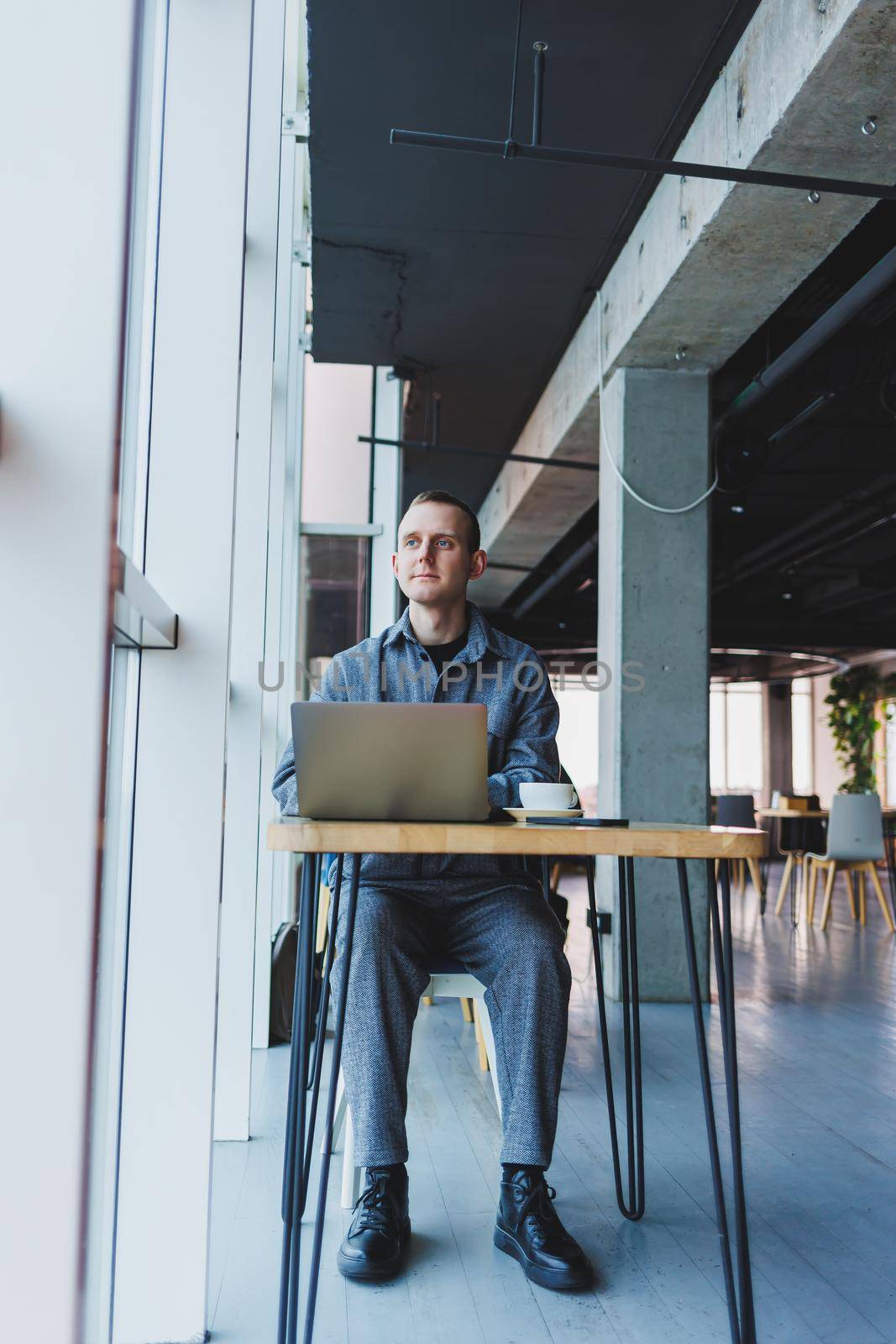 A young successful businessman in a suit drinks coffee and works in a cafe on wireless free internet on a laptop, sitting alone at a table in a cafe.