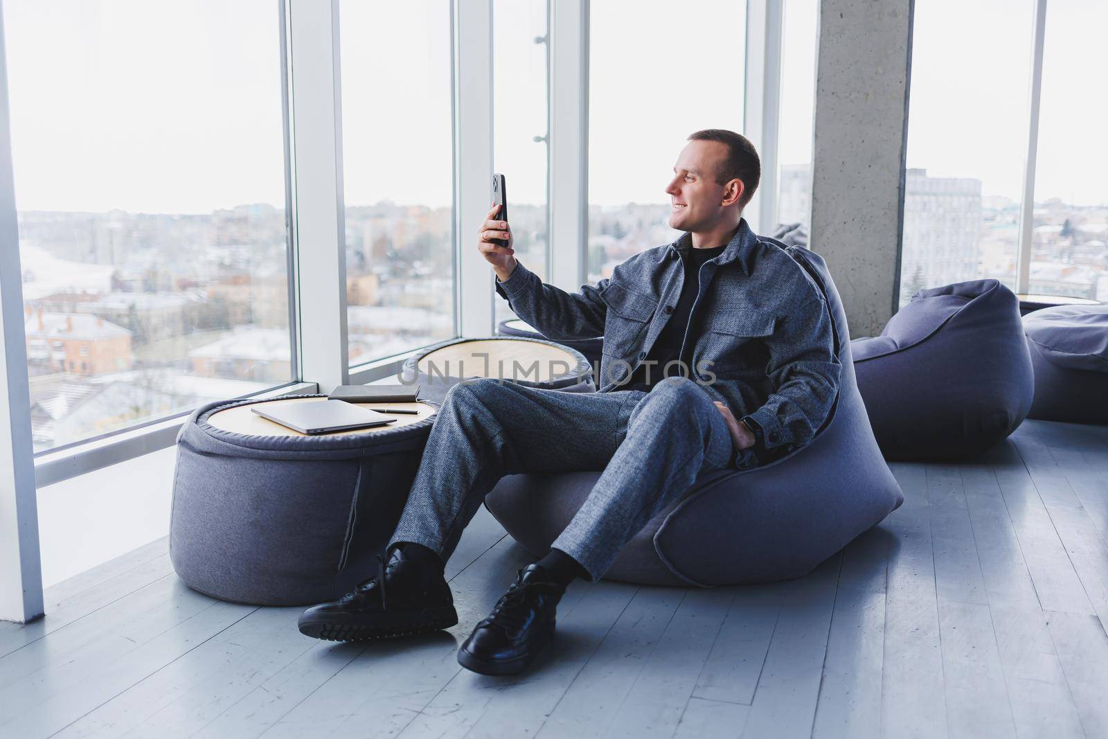 Young male student in a casual suit resting in a comfortable pouf while studying, an attractive freelancer in a suit works remotely