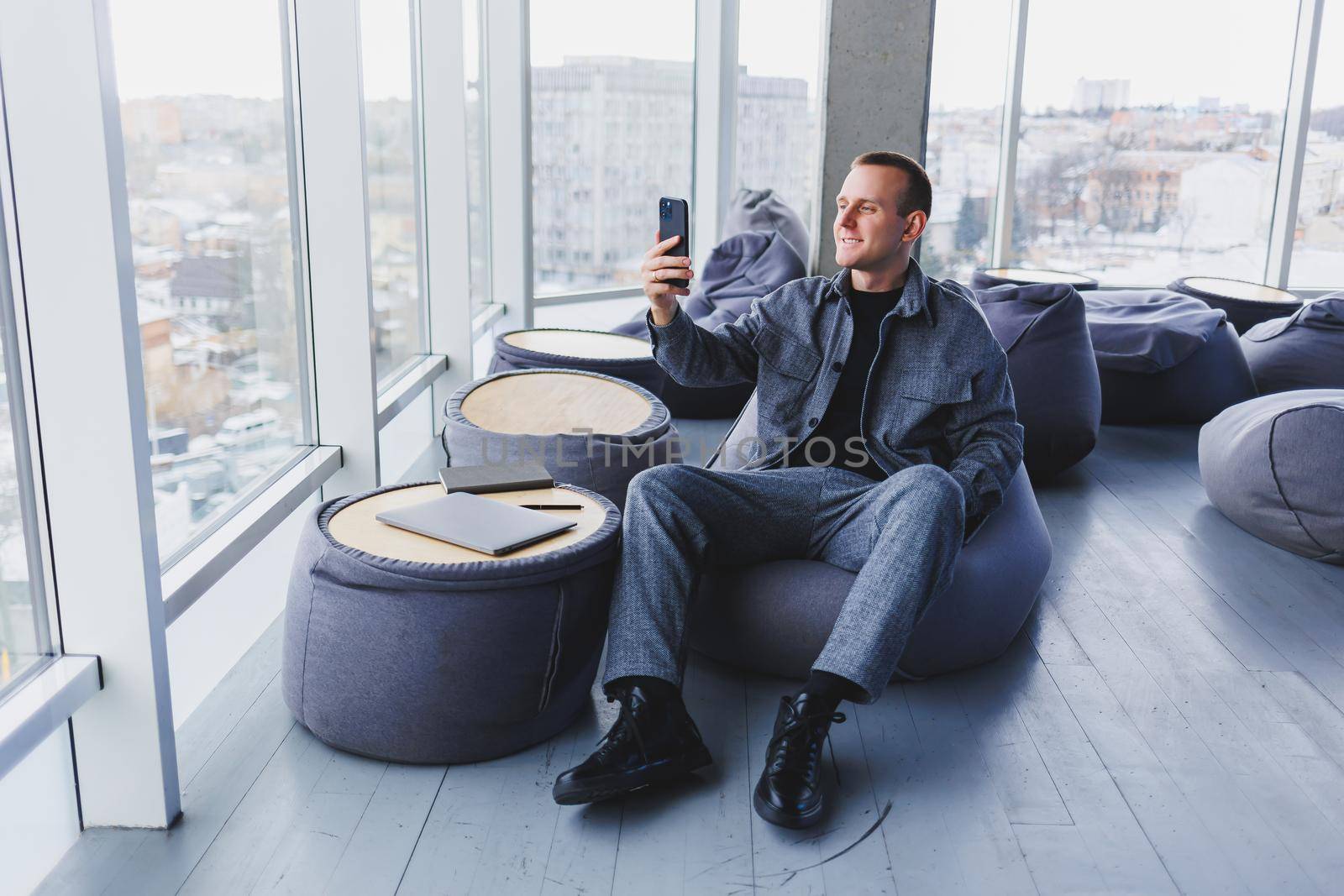 Young male student in a casual suit resting in a comfortable pouf while studying, an attractive freelancer in a suit works remotely