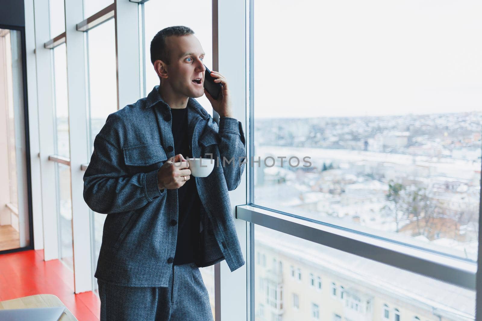 A handsome man of European appearance in a casual suit drinks coffee, talks on the phone and looks out the window