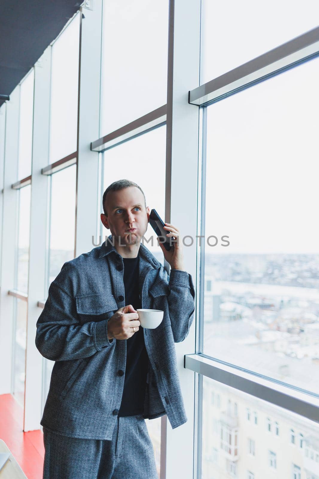A handsome man of European appearance in a casual suit drinks coffee, talks on the phone and looks out the window