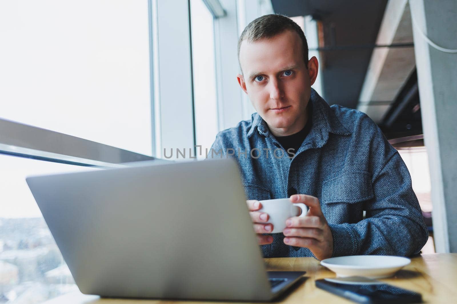 Successful happy business man is sitting at a table in a cafe, holding a cup of coffee and using a laptop.