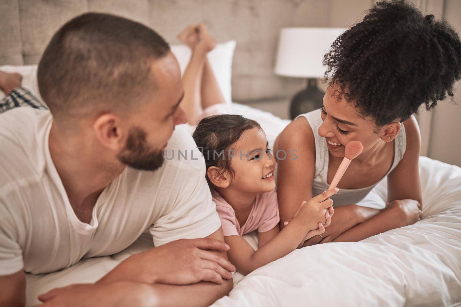 Cosmetics, relax and happy family on a bed with the child having fun with a makeup brush, laughing and playing. Mother, father and girl enjoy quality time together with beauty products in the morning by YuriArcurs