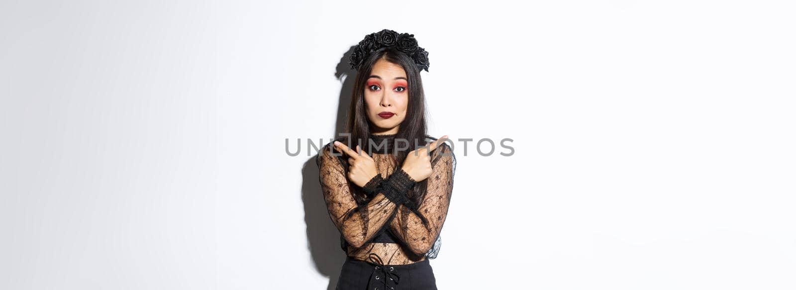 Confused pretty young woman in elegant black lace dress and wreath looking unsure, pointing fingers sideways, showing two variants, halloween banners, standing over white background.