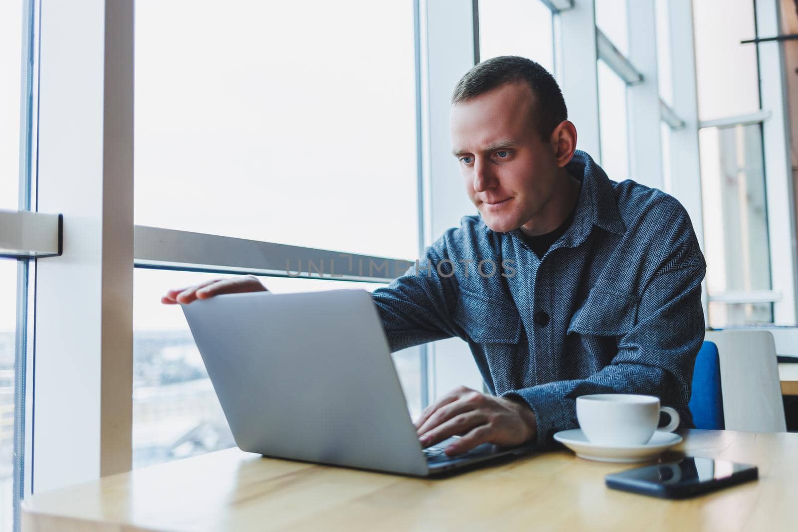 Successful happy business man is sitting at a table in a cafe, holding a cup of coffee and using a laptop.