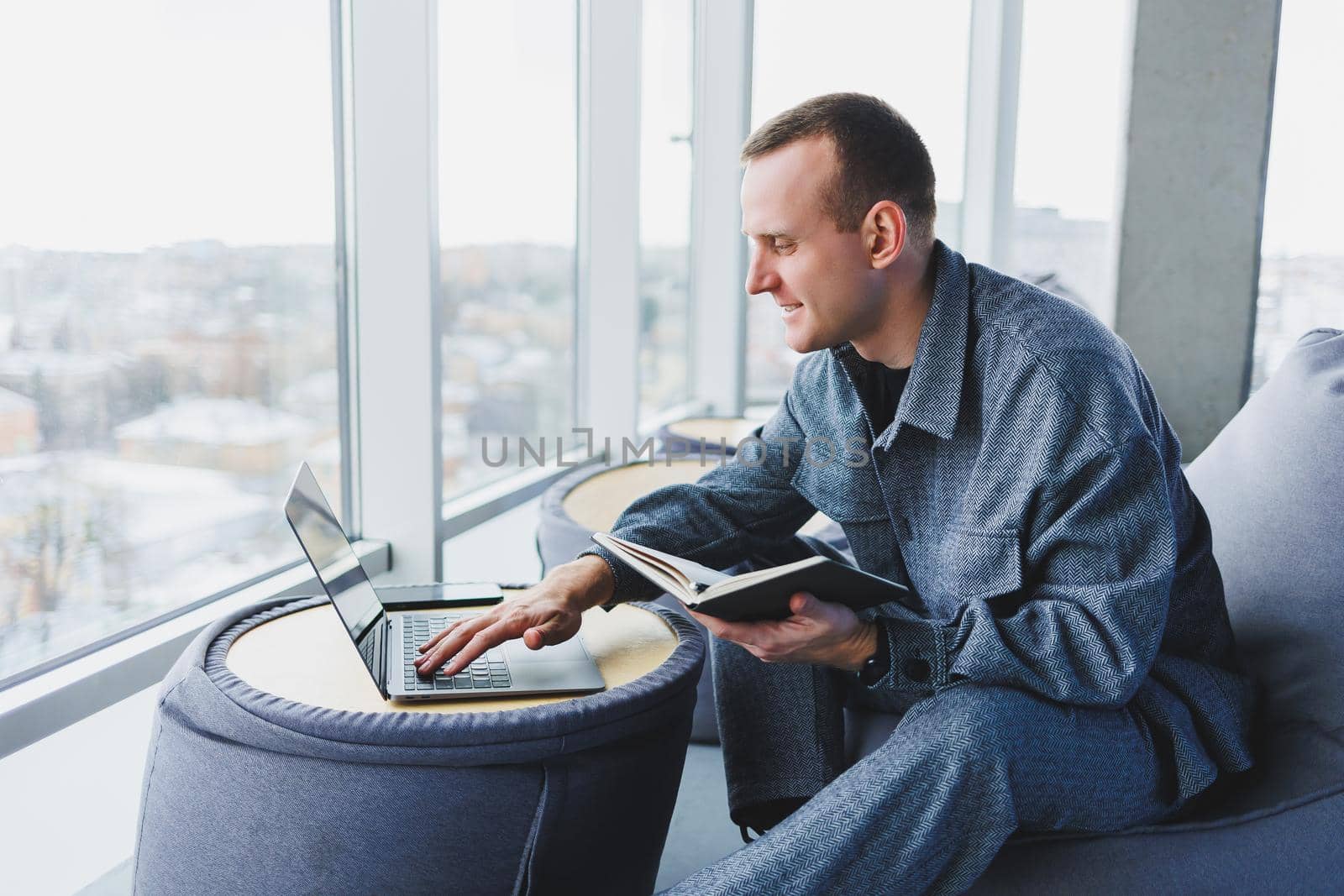 A young male student in a casual suit resting in a comfortable pouf while studying makes notes in a notebook, an attractive freelancer in a suit works remotely