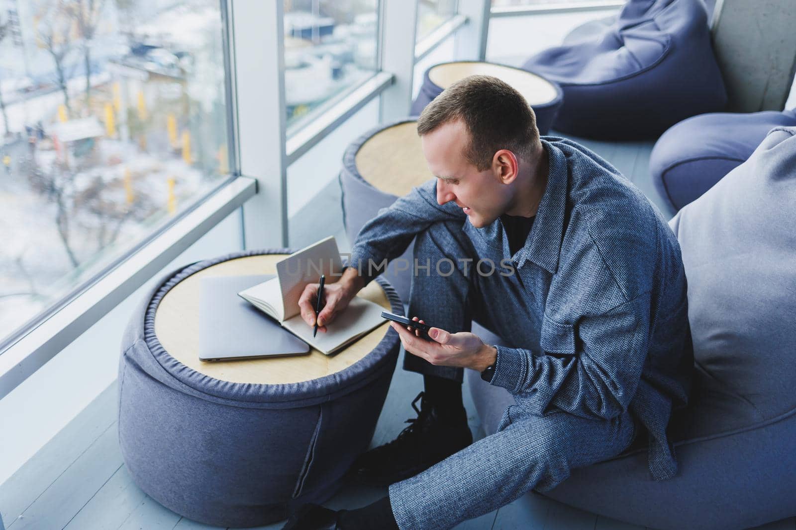 A handsome European young businessman in a coffee shop sits with a mobile phone and makes notes in a notebook. Wearing an elegant casual suit and sitting with a laptop in a public cafe