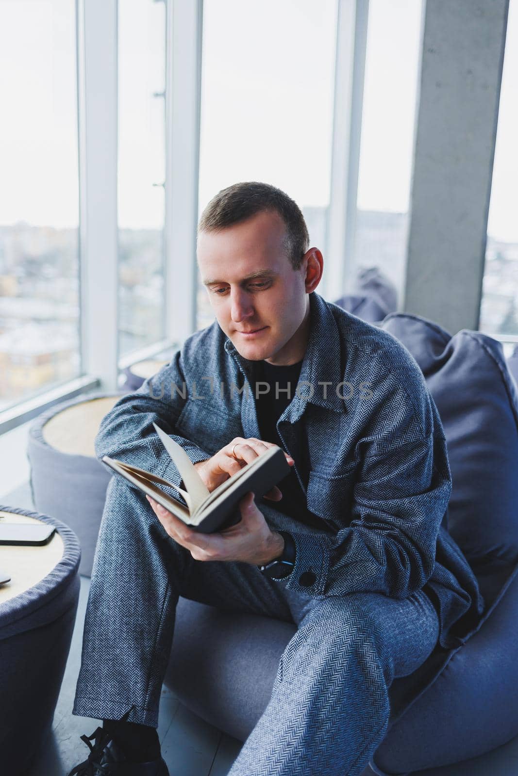 A young male student in a casual suit resting in a comfortable pouf while studying makes notes in a notebook, an attractive freelancer in a suit works remotely