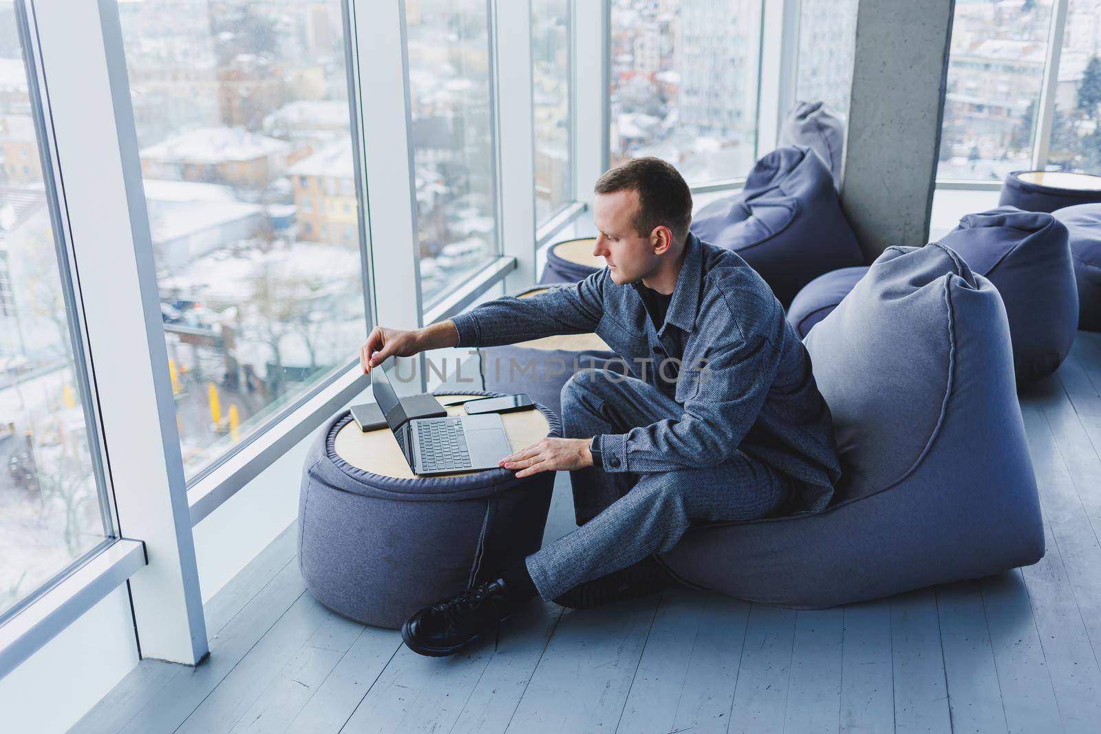 Break. Smiling young man relaxing on bean bag chair while sitting at table and relaxing using laptop, happy businessman leaning back, enjoying work, feeling satisfied
