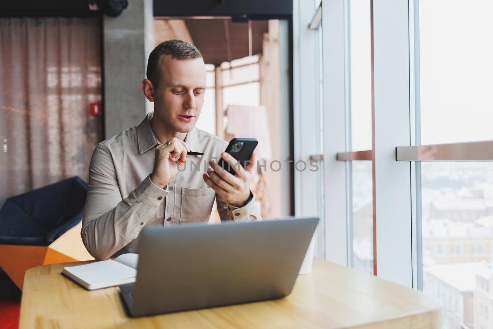 Successful young man is sitting at a table in a cafe-restaurant indoors, working and studying on a laptop computer,holding a mobile phone with a blank screen. Freelancer mobile office business concept