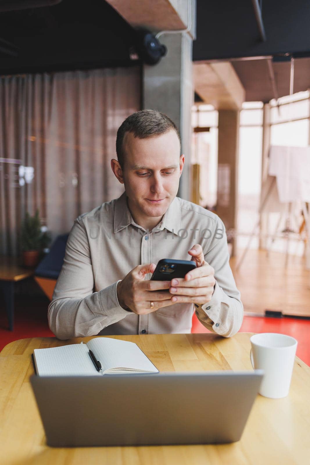 Successful young man is sitting at a table in a cafe-restaurant indoors, working and studying on a laptop computer,holding a mobile phone with a blank screen. Freelancer mobile office business concept