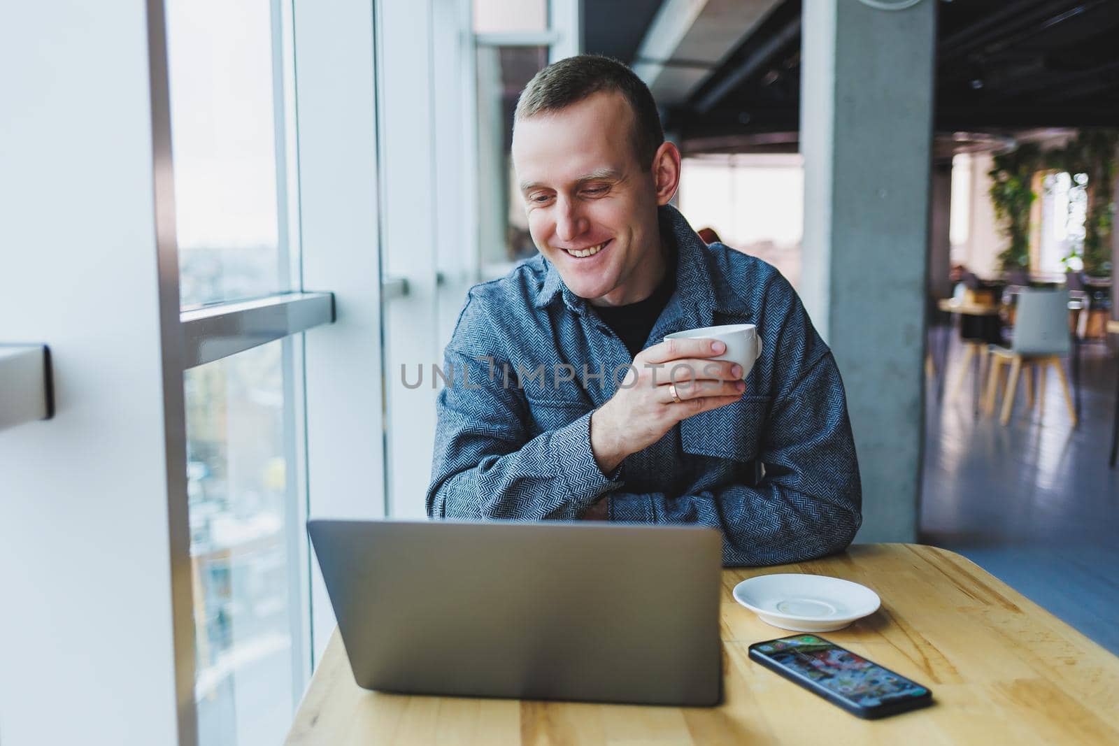 Successful happy business man is sitting at a table in a cafe, holding a cup of coffee and using a laptop.