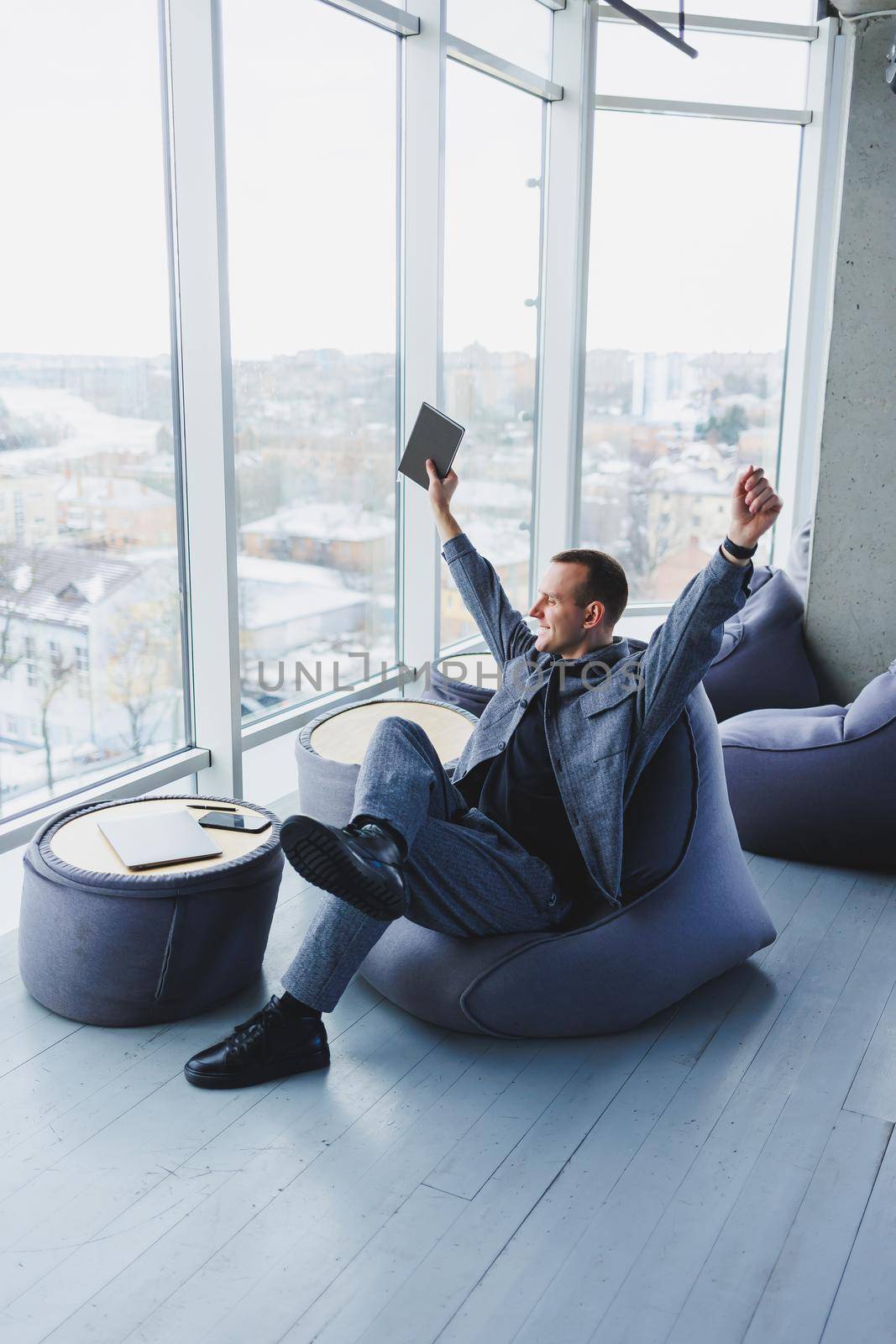 A young male student in a casual suit resting in a comfortable pouf while studying makes notes in a notebook, an attractive freelancer in a suit works remotely