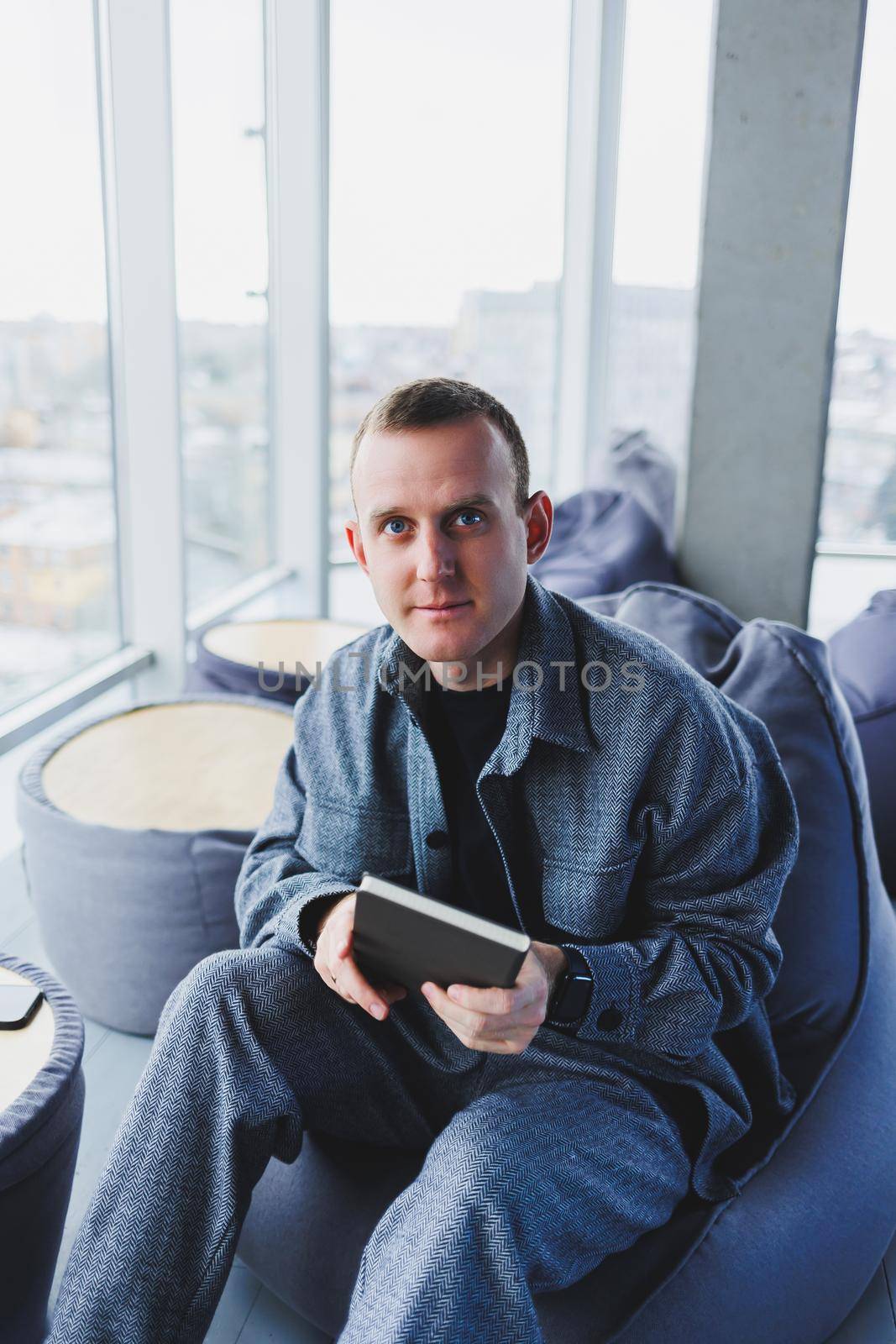 A young male student in a casual suit resting in a comfortable pouf while studying makes notes in a notebook, an attractive freelancer in a suit works remotely