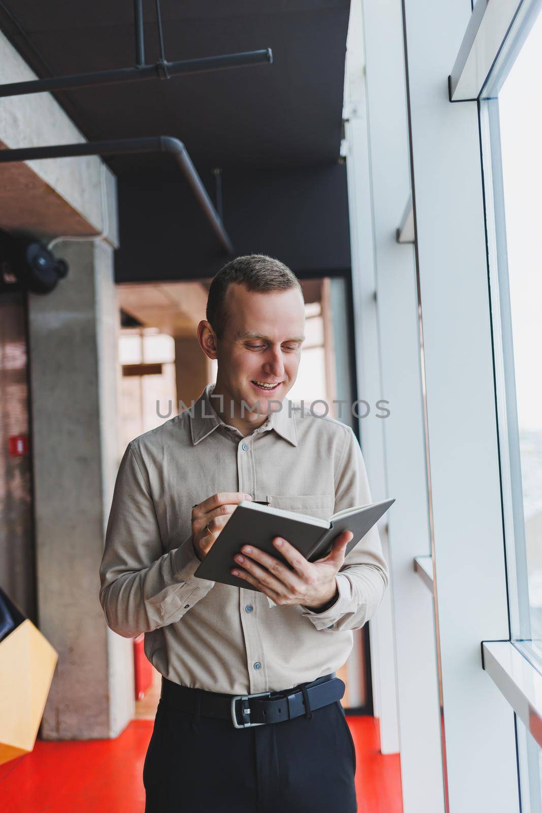 A young businessman stands at a panoramic window in a skyscraper and makes notes in his notebook with a pen. A man in a shirt and trousers of European appearance