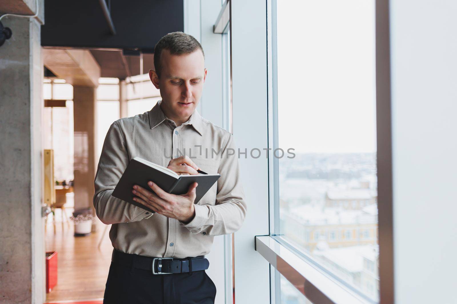 A young businessman stands at a panoramic window in a skyscraper and makes notes in his notebook with a pen. A man in a shirt and trousers of European appearance