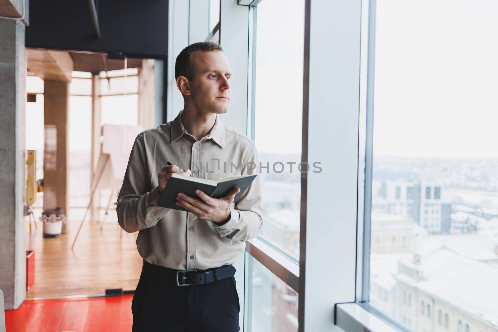 A young businessman stands at a panoramic window in a skyscraper and makes notes in his notebook with a pen. A man in a shirt and trousers of European appearance