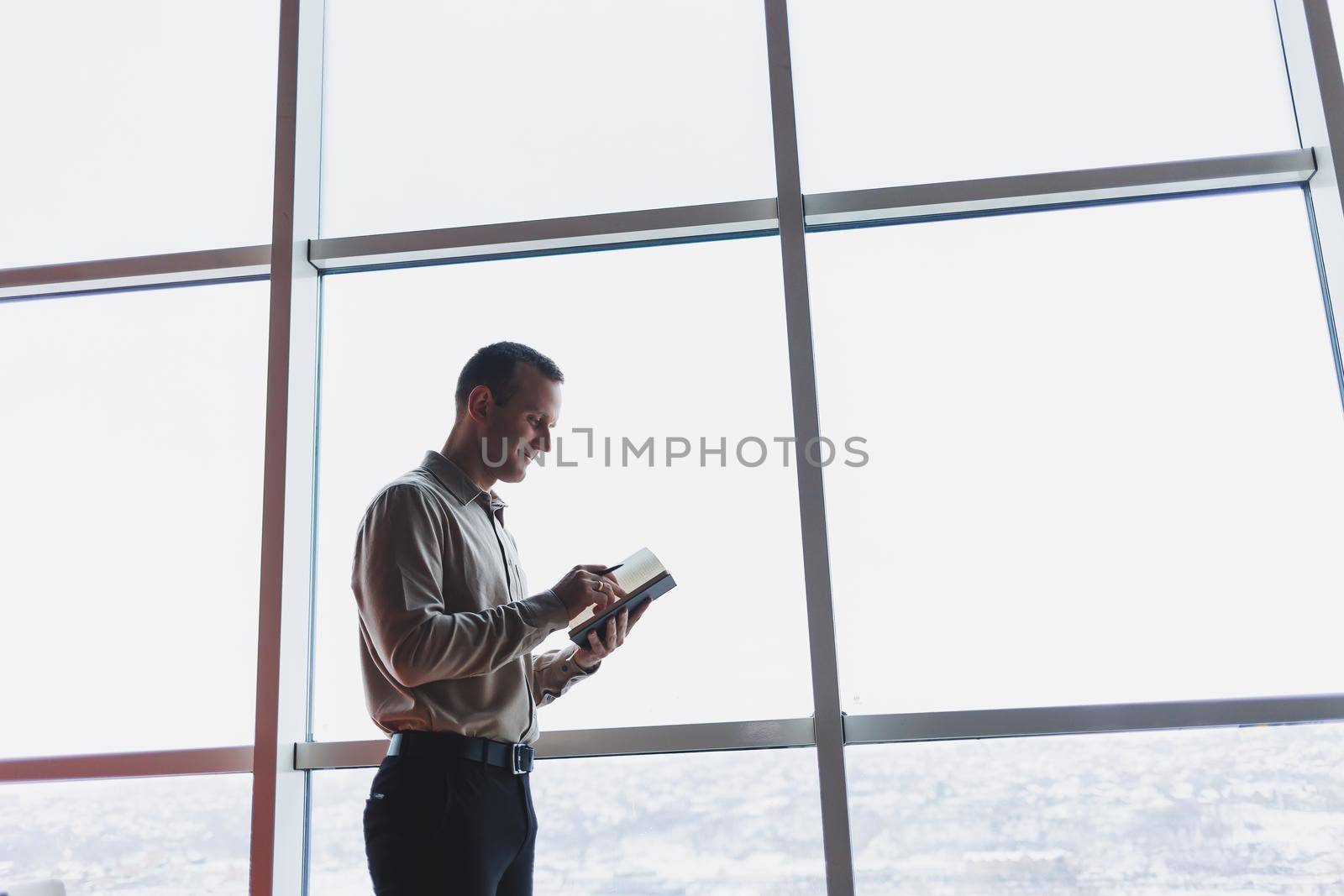 A young businessman stands at a panoramic window in a skyscraper and makes notes in his notebook with a pen. A man in a shirt and trousers of European appearance