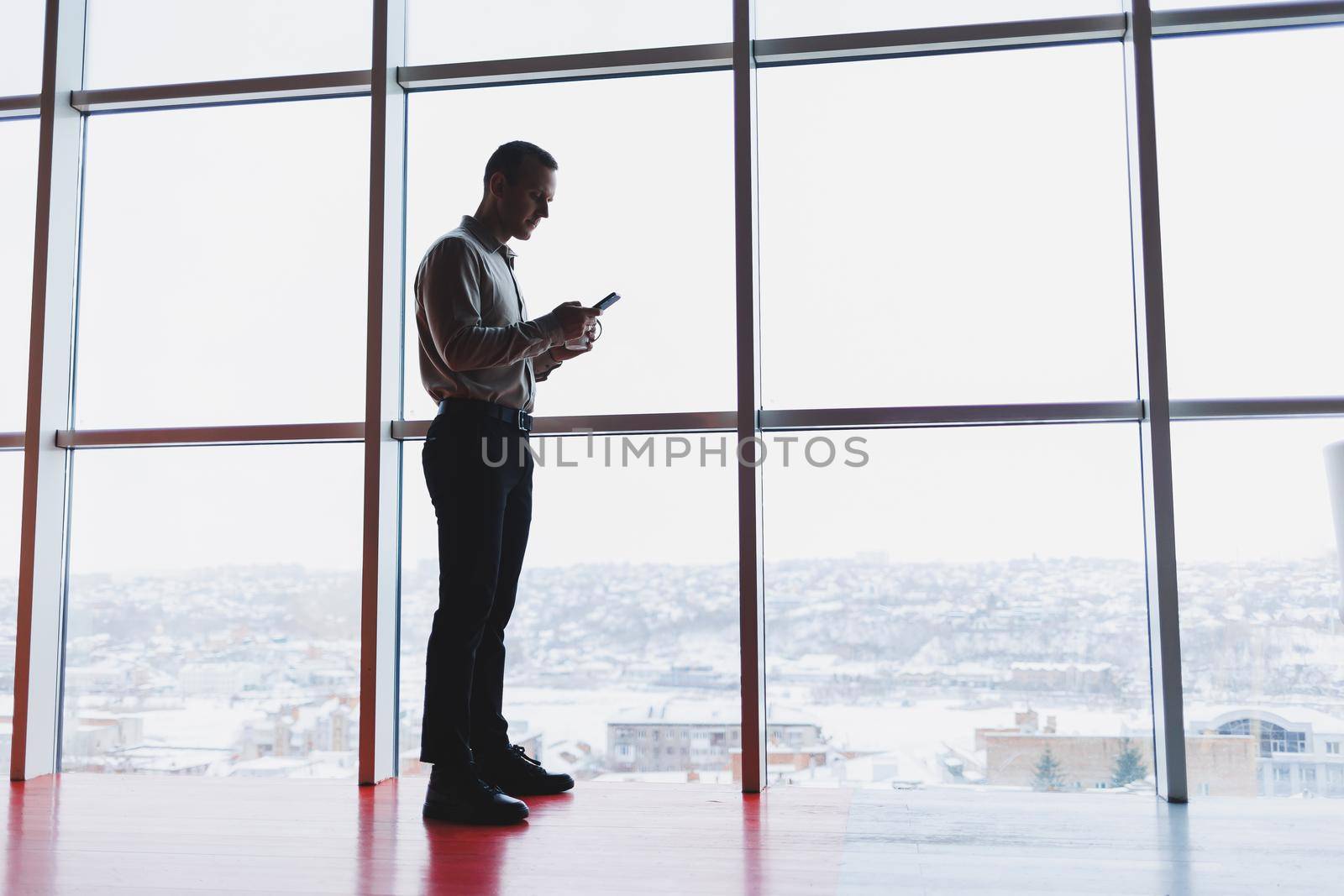 A young executive is holding a phone while standing in an office interior and looking out a large window overlooking the city. Male manager with a phone is thinking about a new business project