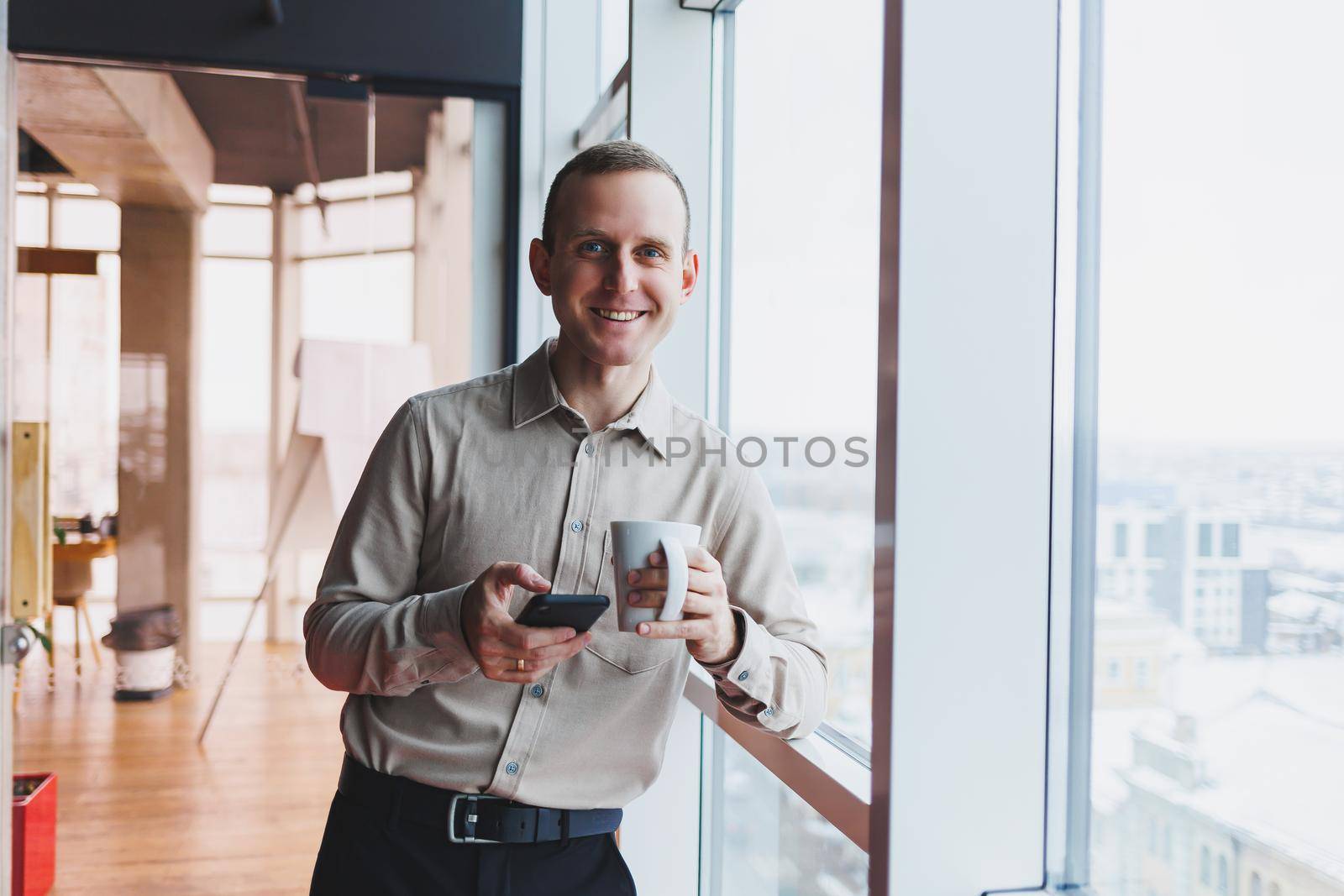 Male brokers with phone looking towards window with coffee in hand in modern workspace