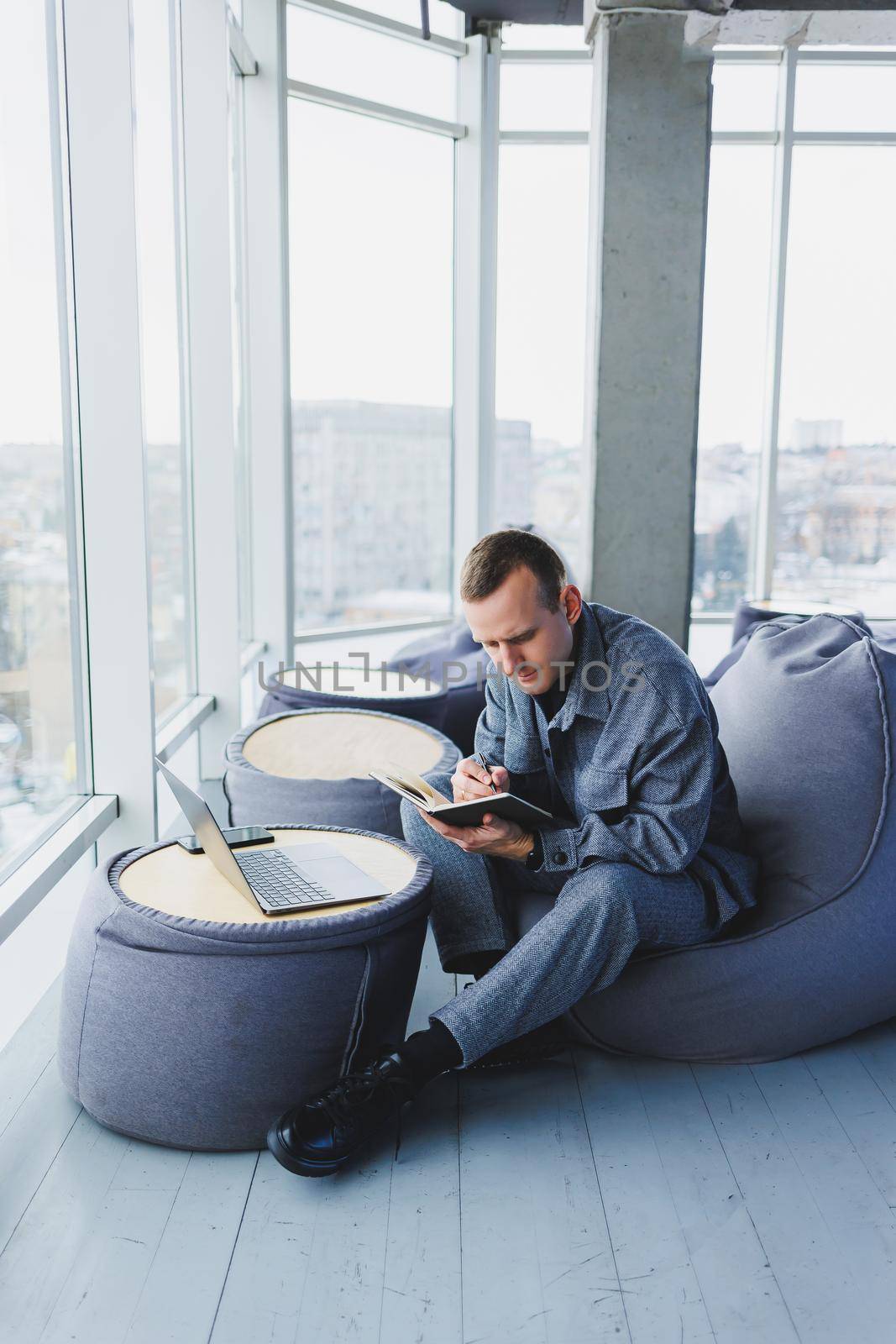 A young male student in a casual suit resting in a comfortable pouf while studying makes notes in a notebook, an attractive freelancer in a suit works remotely