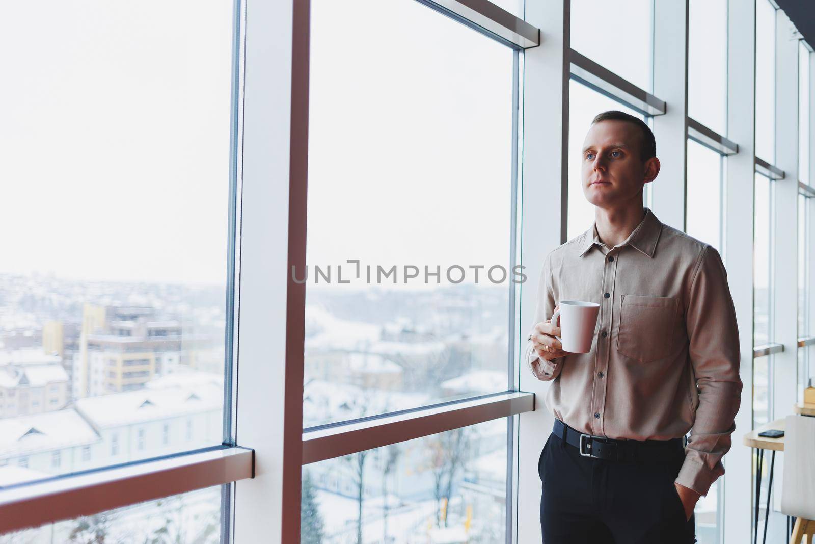 Men in shirt and trousers looking towards window with coffee in hand, in modern workspace