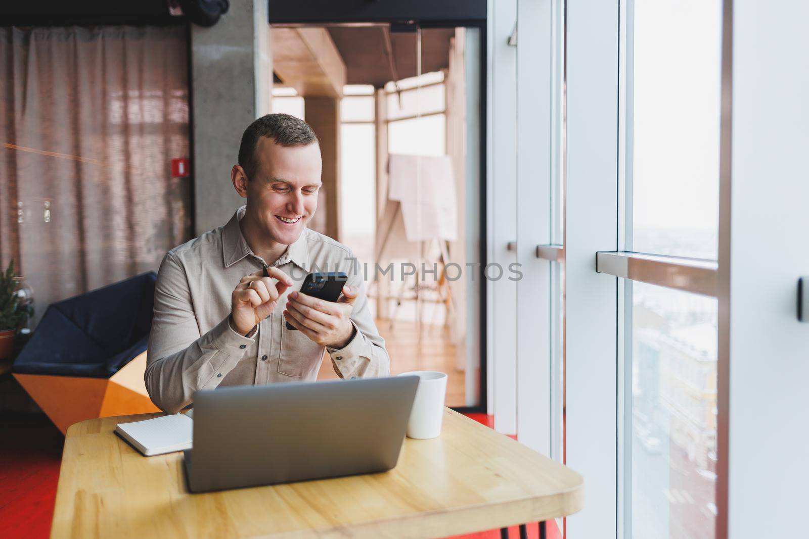 Successful young man is sitting at a table in a cafe-restaurant indoors, working and studying on a laptop computer,holding a mobile phone with a blank screen. Freelancer mobile office business concept