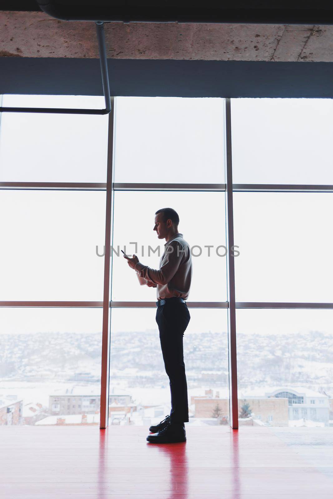 A young executive is holding a phone while standing in an office interior and looking out a large window overlooking the city. Male manager with a phone is thinking about a new business project