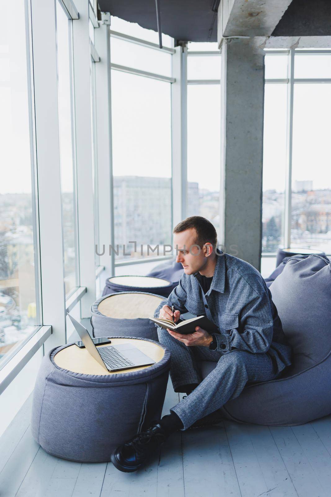 A young male student in a casual suit resting in a comfortable pouf while studying makes notes in a notebook, an attractive freelancer in a suit works remotely