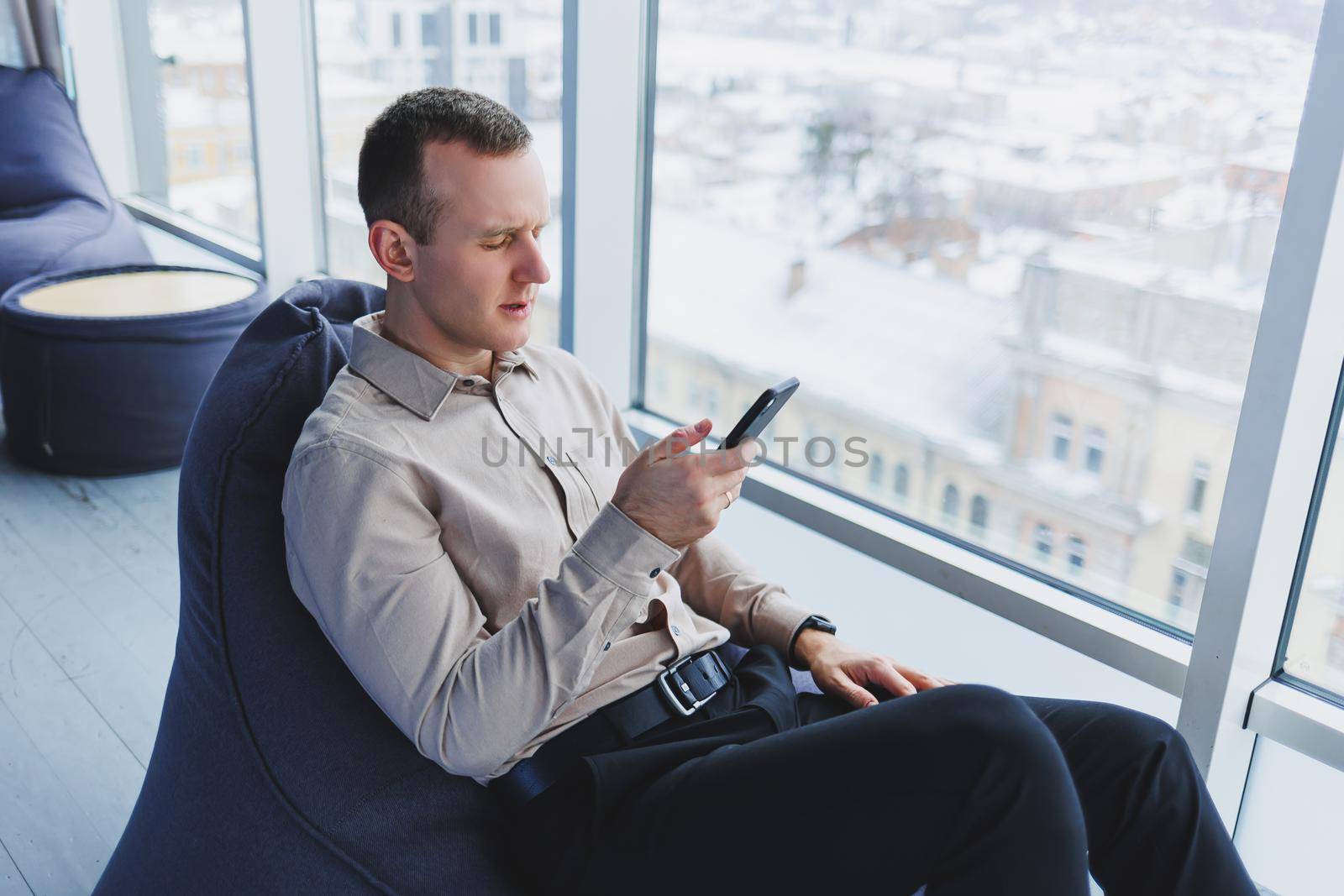 Successful male freelancer sitting in modern workspace and checking notifications on mobile phone in casual clothes. Remote work.