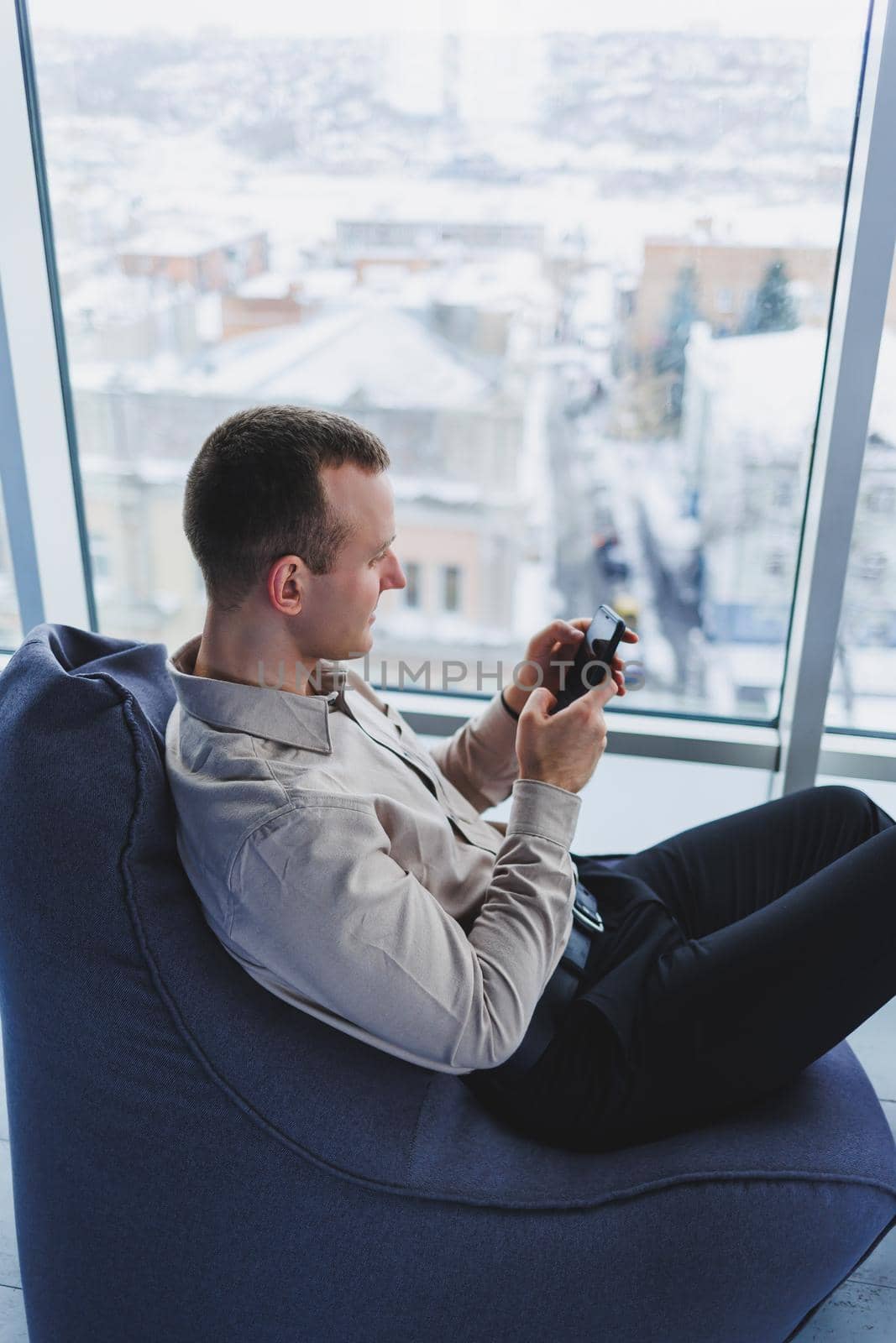 Successful male businessman sitting in a modern workspace and checking notifications on his mobile phone, he is wearing a shirt and trousers. Remote work.