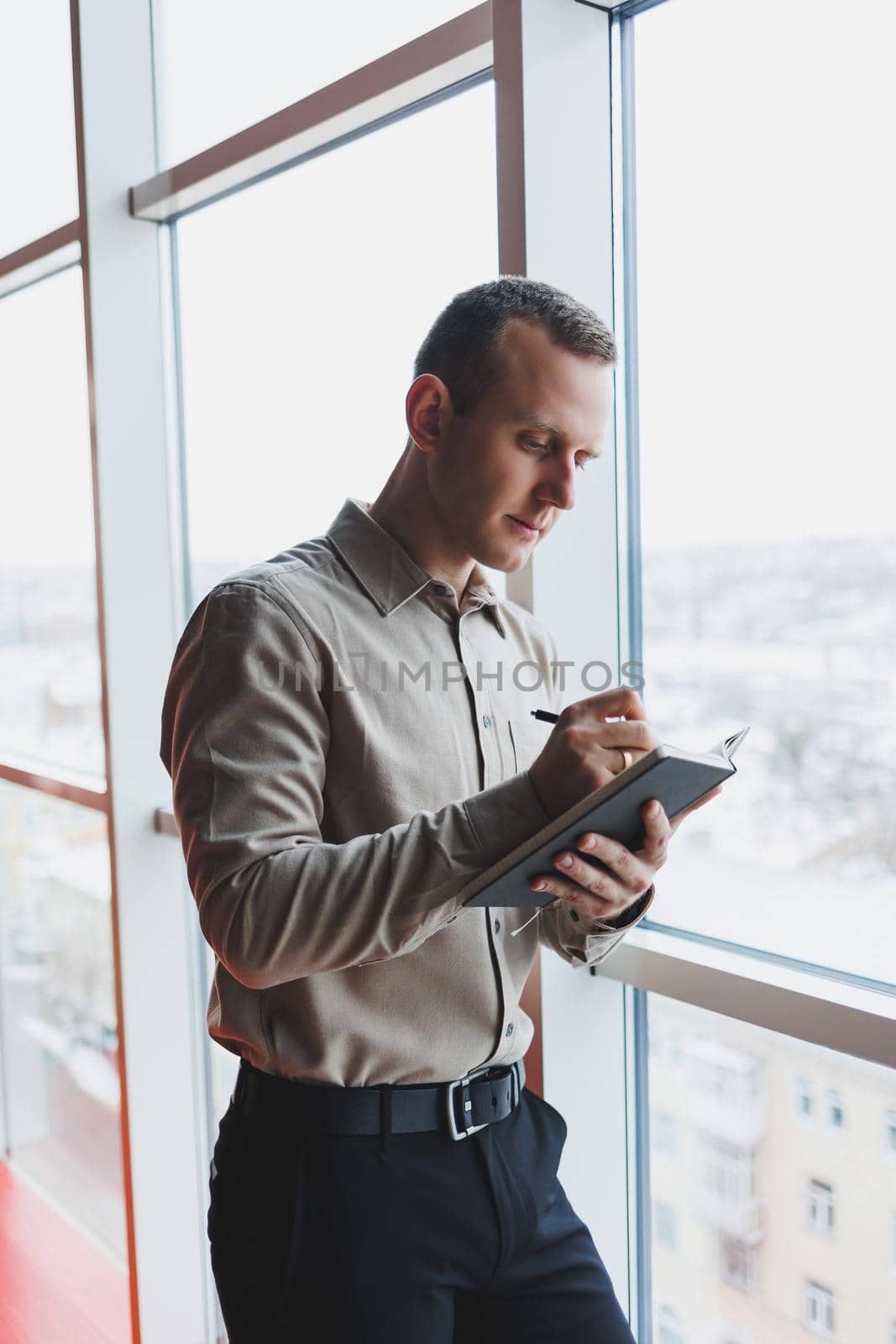 A young businessman stands at a panoramic window in a skyscraper and makes notes in his notebook with a pen. A man in a shirt and trousers of European appearance